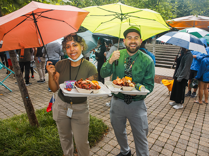 Two Tulane staffers hold umbrellas and plates of boiled seafood and smile at the camera during Juneteenth on Tulane's Uptown campus.