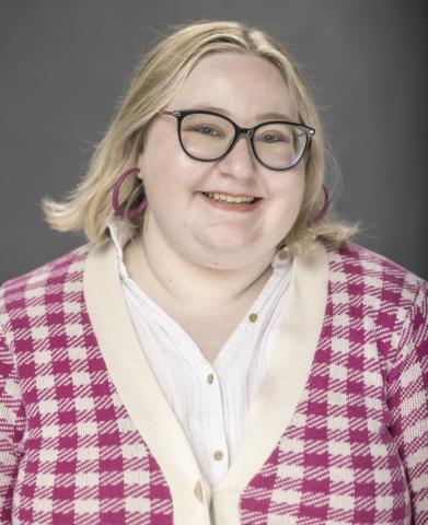 A smiling white person with blonde hair and glasses, wearing a pink and white checkered cardigan.