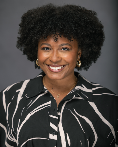 A smiling Black woman with an afro hairstyle poses in a black and beige patterned blouse.